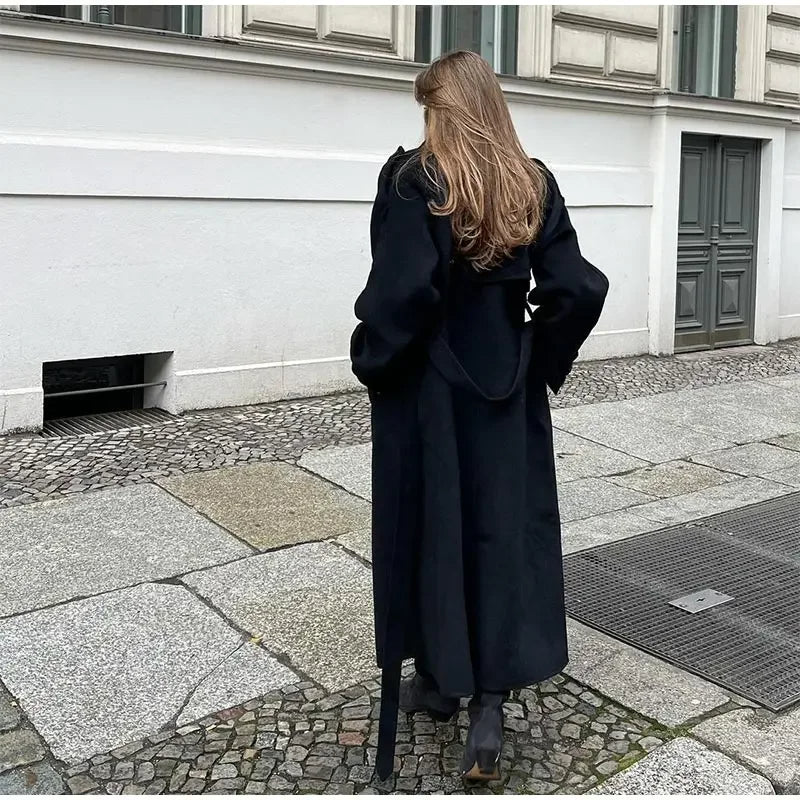 Woman wearing a black longline double-breasted trench coat with wide lapels and a waist belt