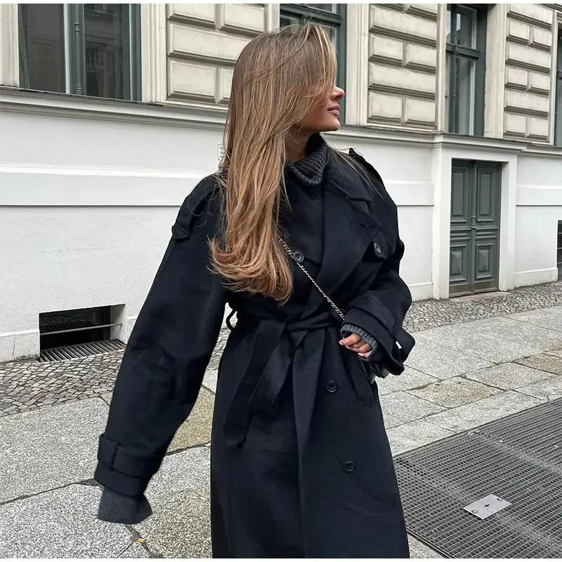 Woman wearing a black longline double-breasted trench coat with wide lapels and a waist belt