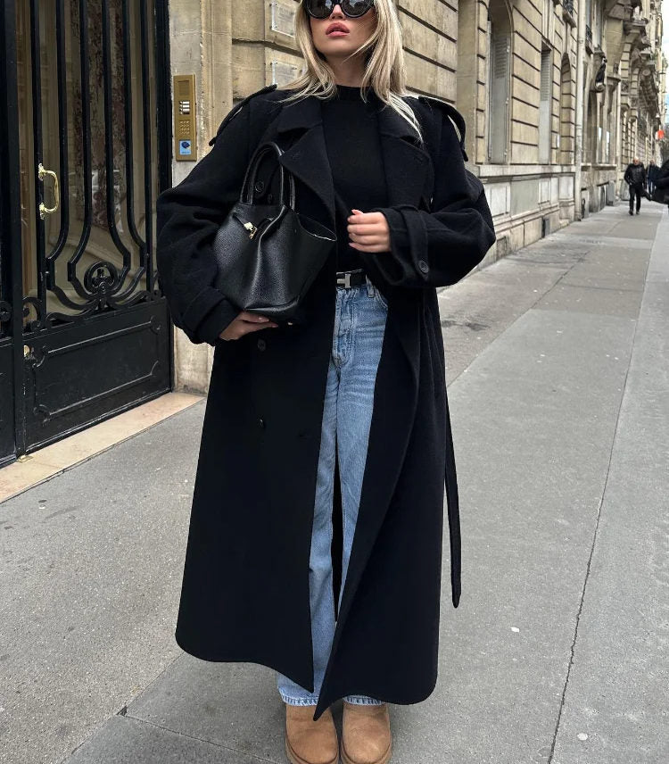 Woman wearing a black longline double-breasted trench coat with wide lapels and a waist belt