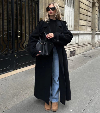 Woman wearing a black longline double-breasted trench coat with wide lapels and a waist belt